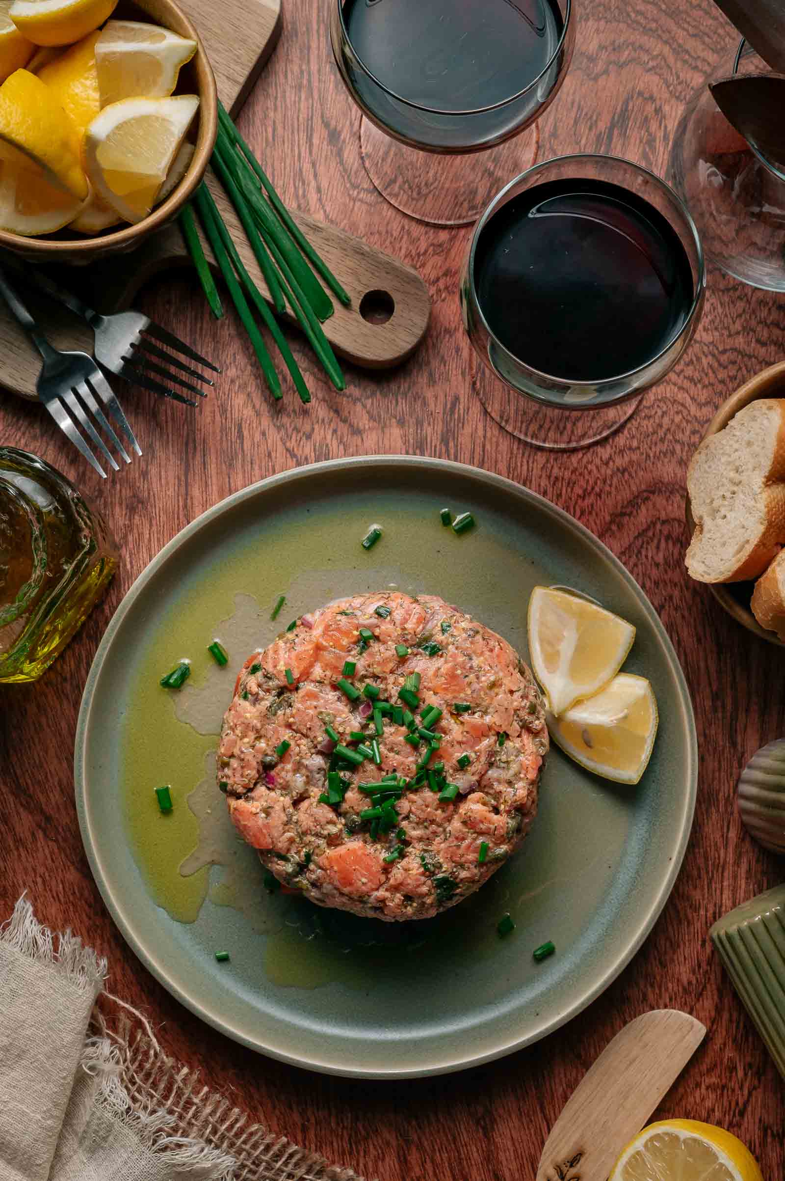 Prato com tártaro de salmão guarnecido com cebolinha picada e rodelas de limão, rodeado de taças de vinho, rodelas de pão, limão e utensílios sobre mesa de madeira.