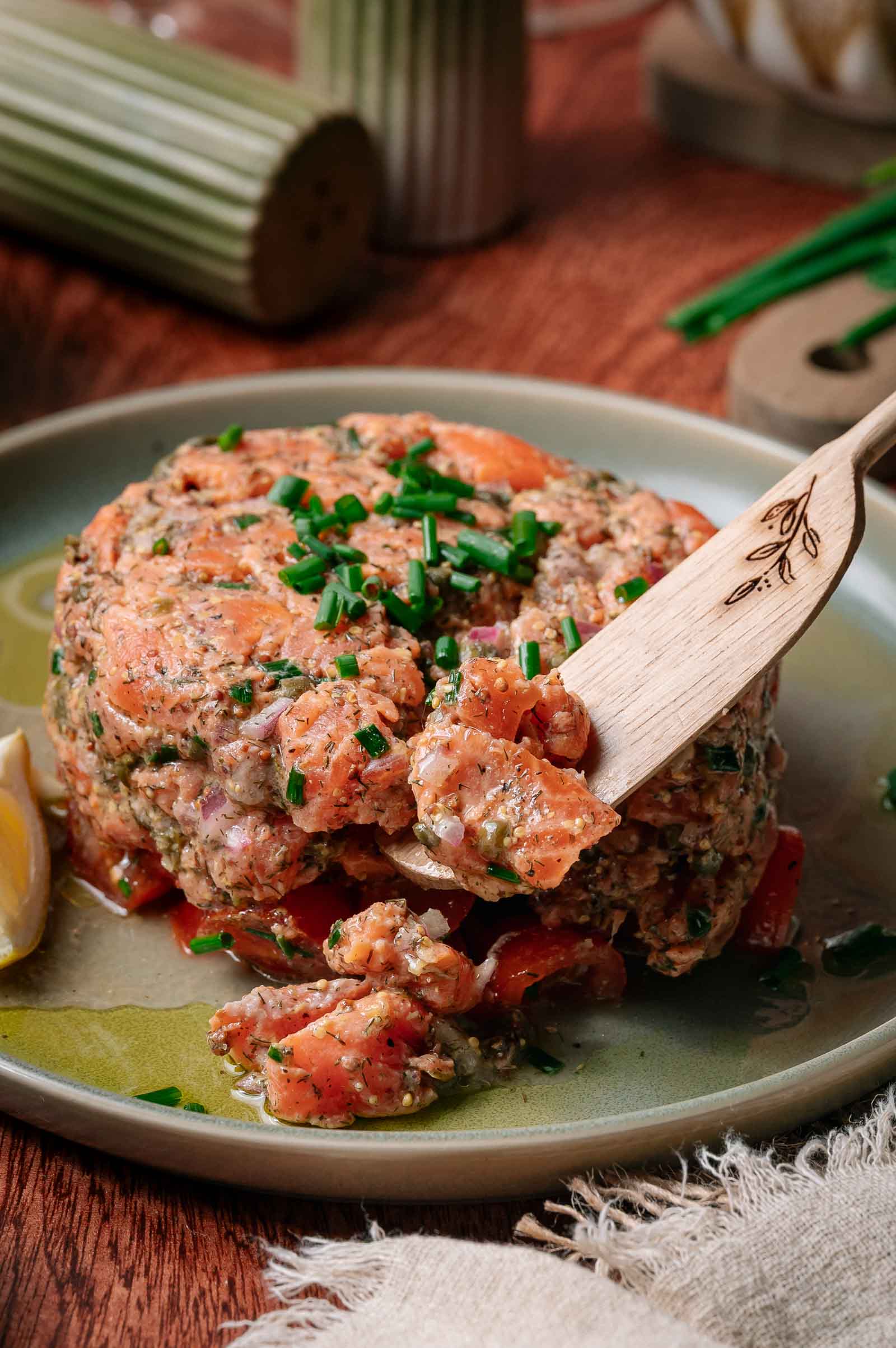 Close de uma porção de tártaro de salmão com ervas e legumes em cubos, guarnecido com cebolinha picada em um prato verde, com um garfo de madeira e uma rodela de limão ao lado.