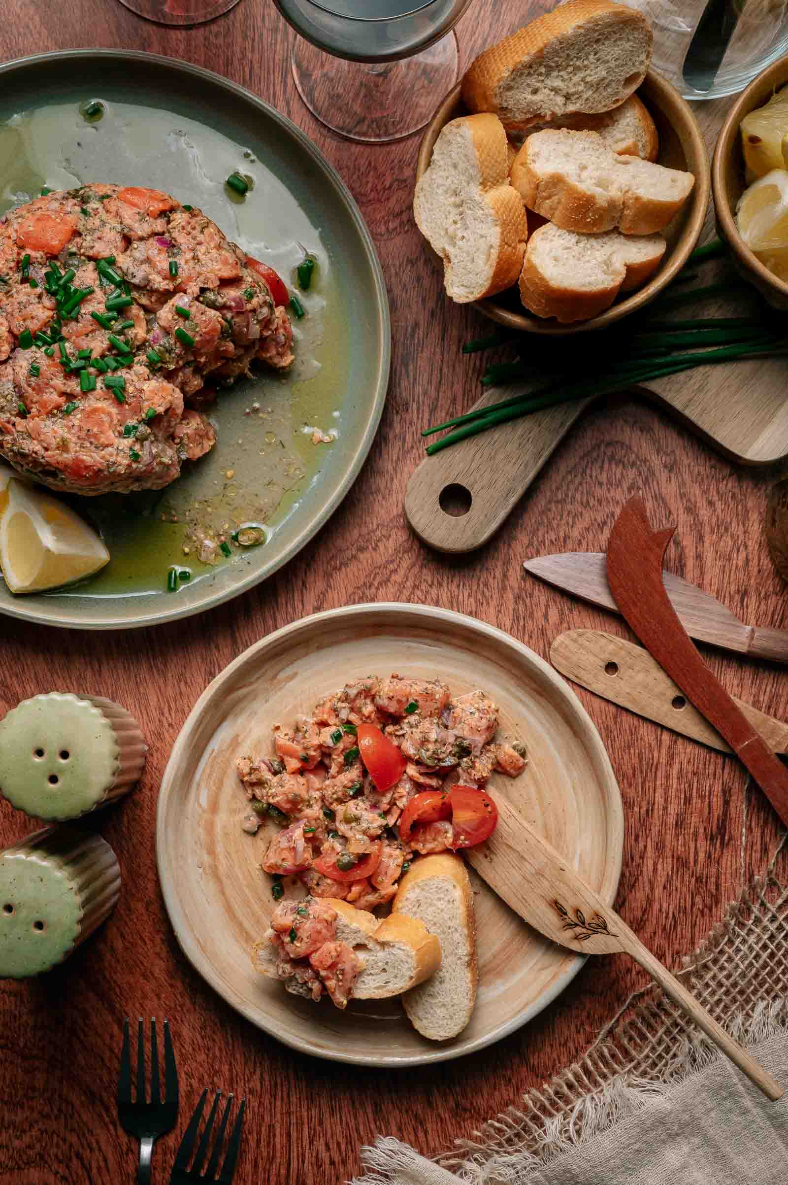 Dois pratos de tártaro de salmão com tomate picado e ervas, servidos com rodelas de limão e baguete fatiada sobre mesa de madeira posta com utensílios e guarnições.