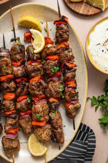 Espetos de cordeiro grelhado e vegetais com pimentão vermelho e cebola em um prato, guarnecidos com rodelas de limão e ervas frescas, ao lado de uma tigela de molho tzatziki branco.