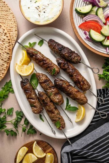 Kofta de cordeiro grelhado em espetos de metal servido em prato oval branco com rodelas de limão e folhas de hortelã, rodeado de pão pita, molho tzatziki e salada de pepino e tomate.