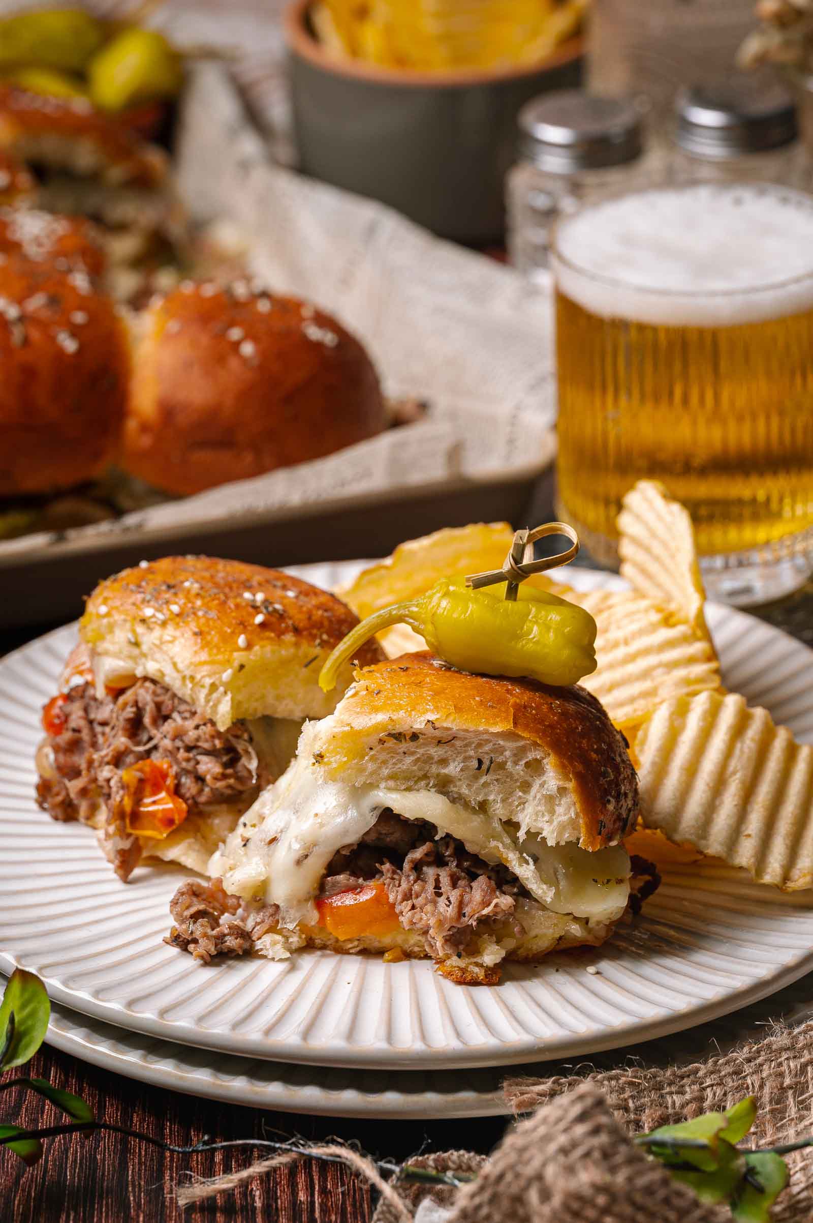 Dois controles deslizantes de cheesesteak philly em um prato com batatas fritas e pimenta em conserva, com um copo de cerveja e mais controles deslizantes ao fundo.