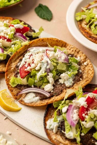 Gyro smashburgers cobertos com cordeiro moído, alface, tomate, cebola roxa, queijo feta e molho branco, servidos com rodelas de limão.
