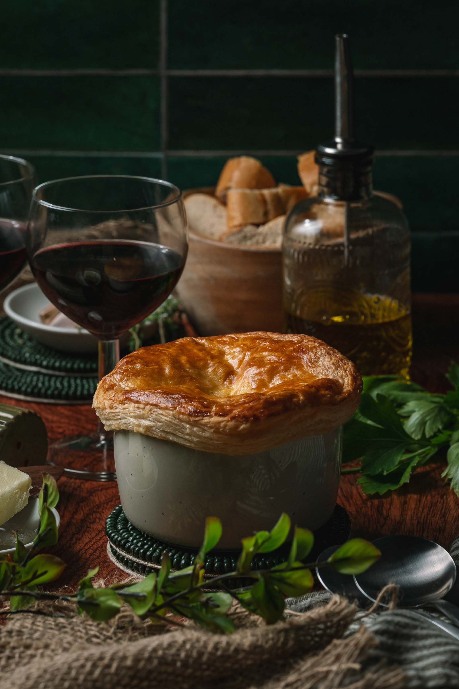 Uma torta de lagosta com massa folhada marrom-dourada em um ramekin branco fica sobre uma mesa ao lado de uma taça de vinho tinto, pão, azeite e ervas frescas.