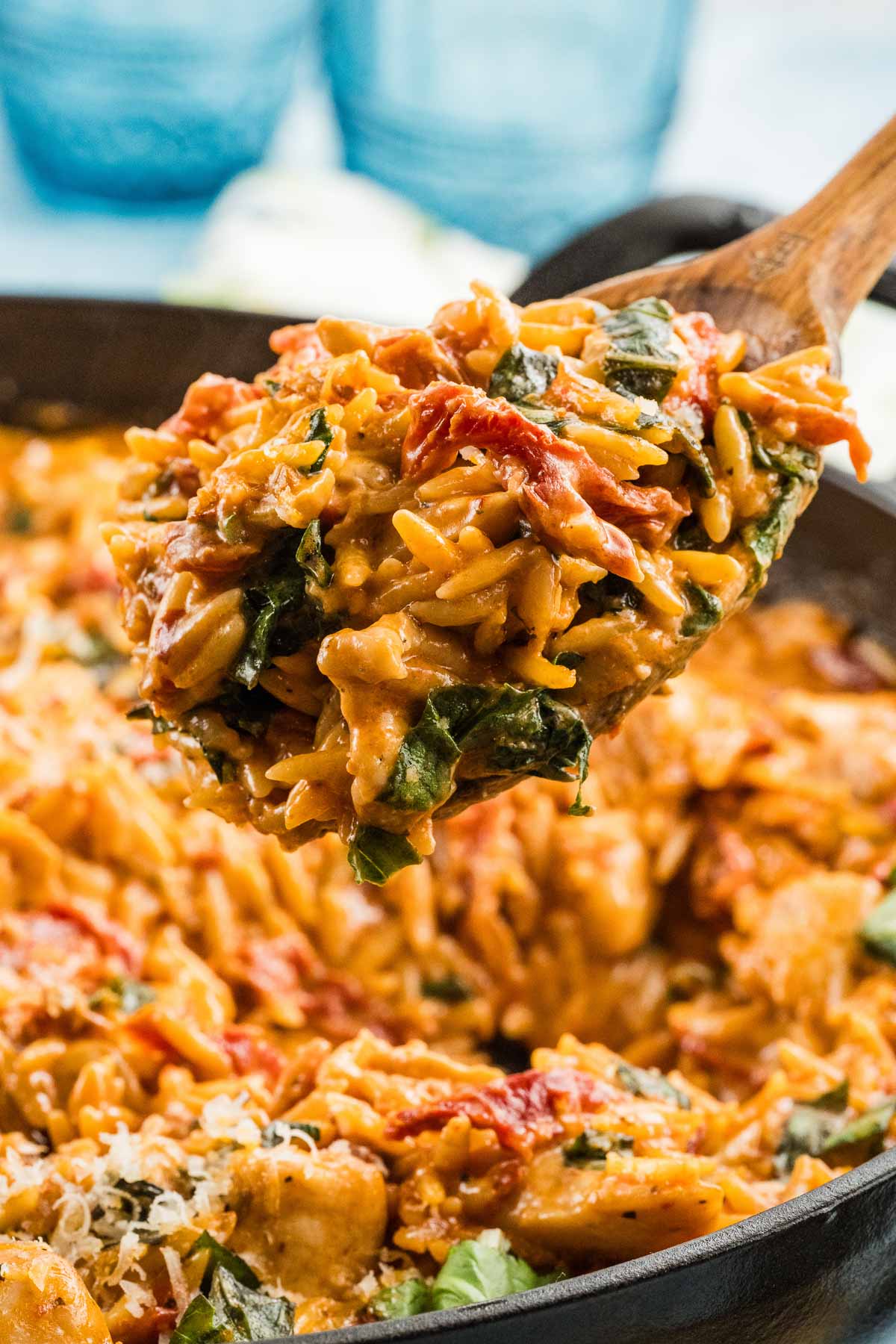 Uma colher de pau levanta uma porção de frango orzo cremoso case comigo com manjericão, tomate seco e frango de uma frigideira.