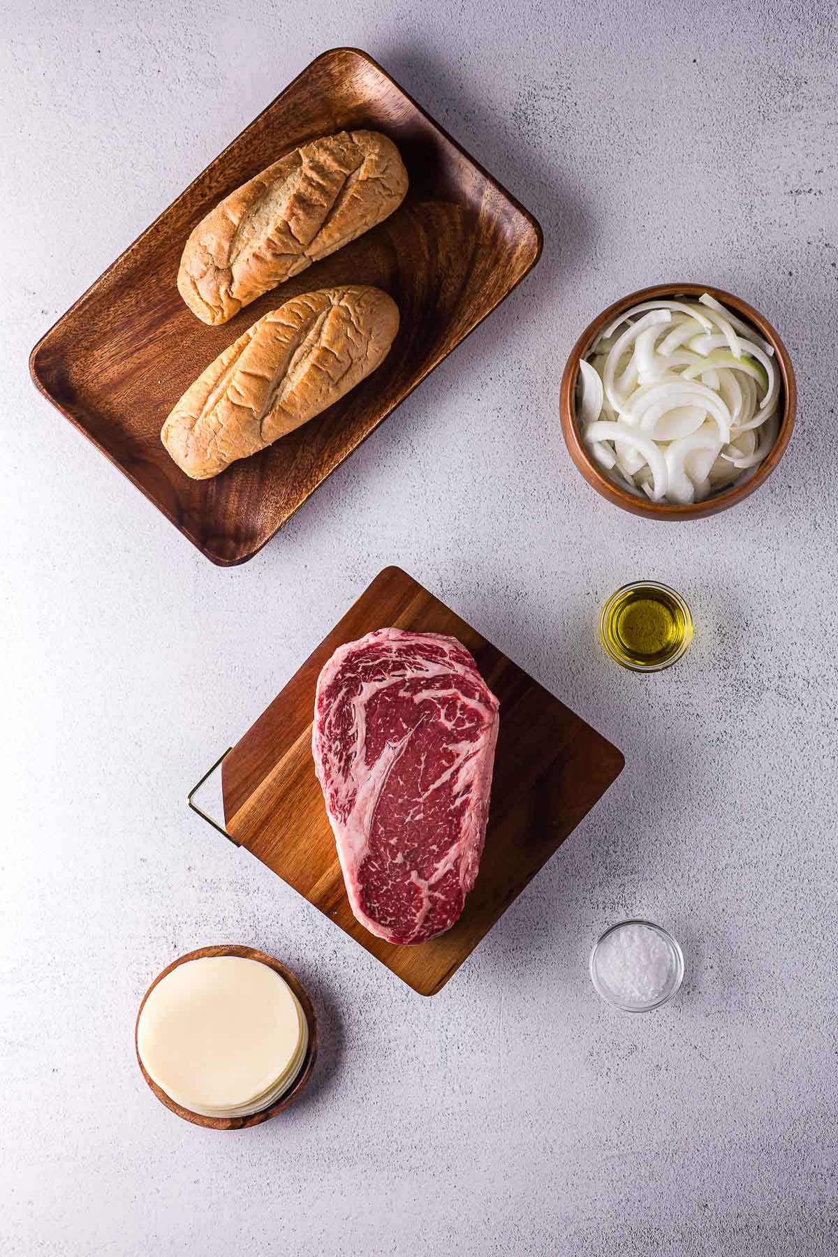 Um bife, dois pãezinhos, cebolas fatiadas, uma tigela pequena de azeite, uma tigela de sal e queijo fatiado estão dispostos sobre uma superfície branca.