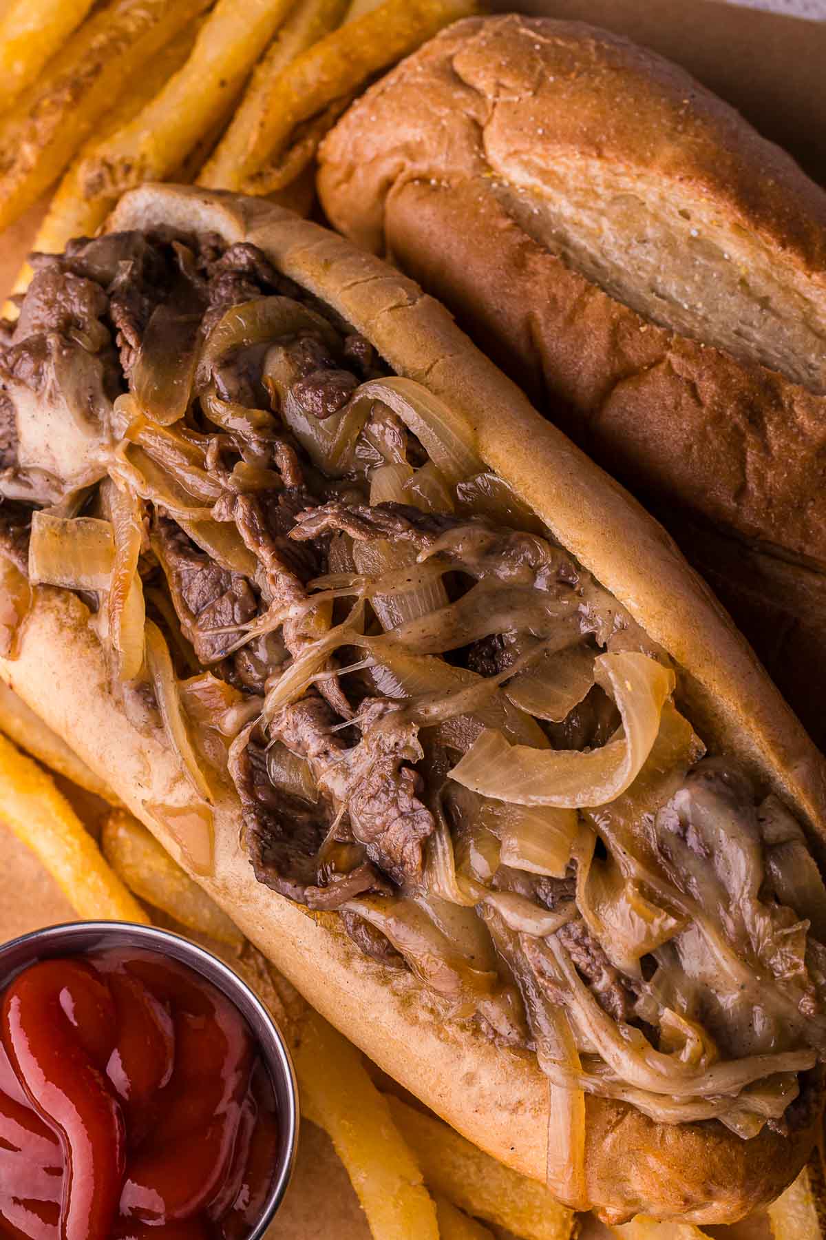 Sanduíche de cheesesteak Philly com cebola salteada, servido com batatas fritas, acompanhamento de ketchup e pãozinho hoagie.