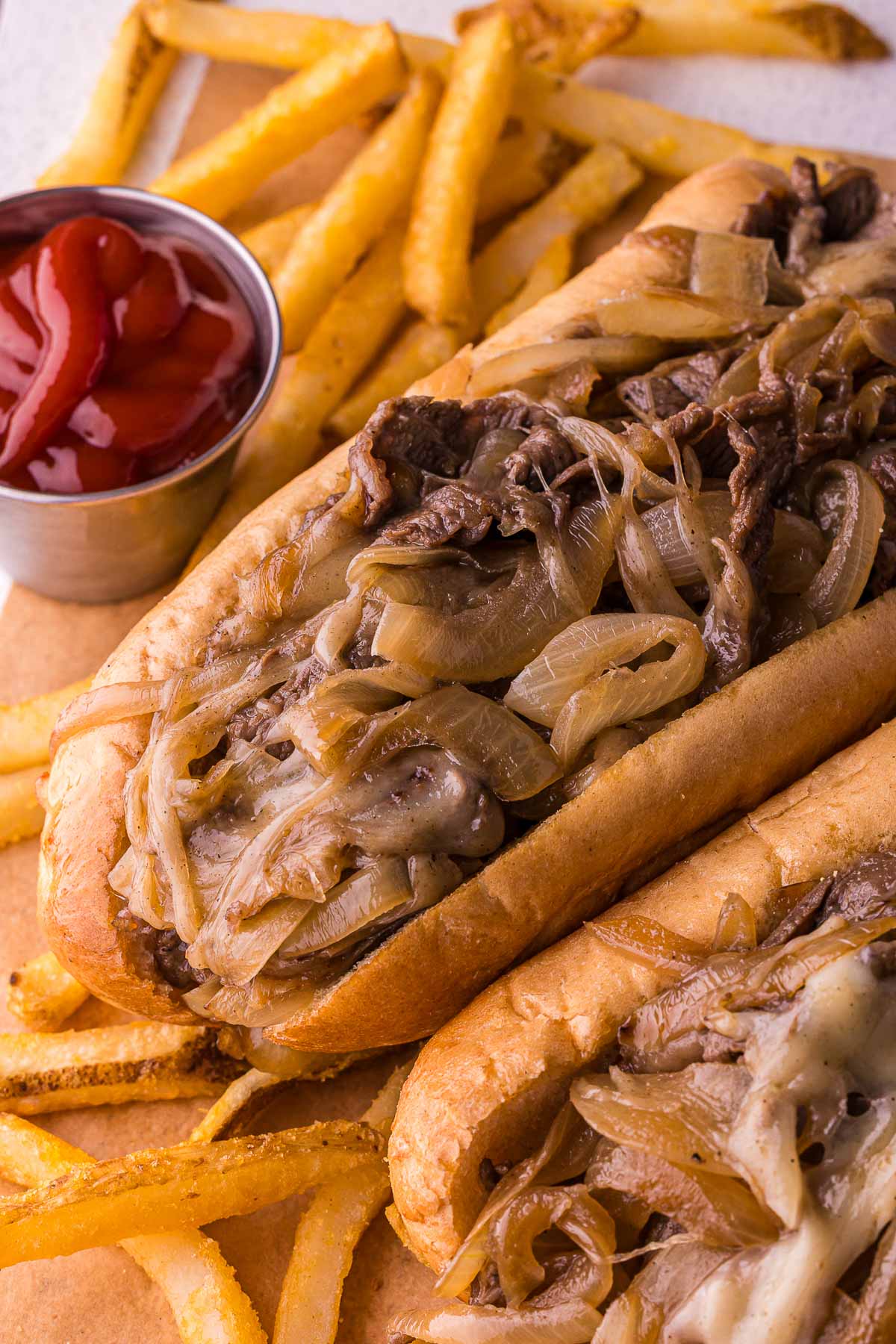 Um close de dois cheesesteaks com cebola e um acompanhamento de batatas fritas e uma pequena xícara de ketchup.