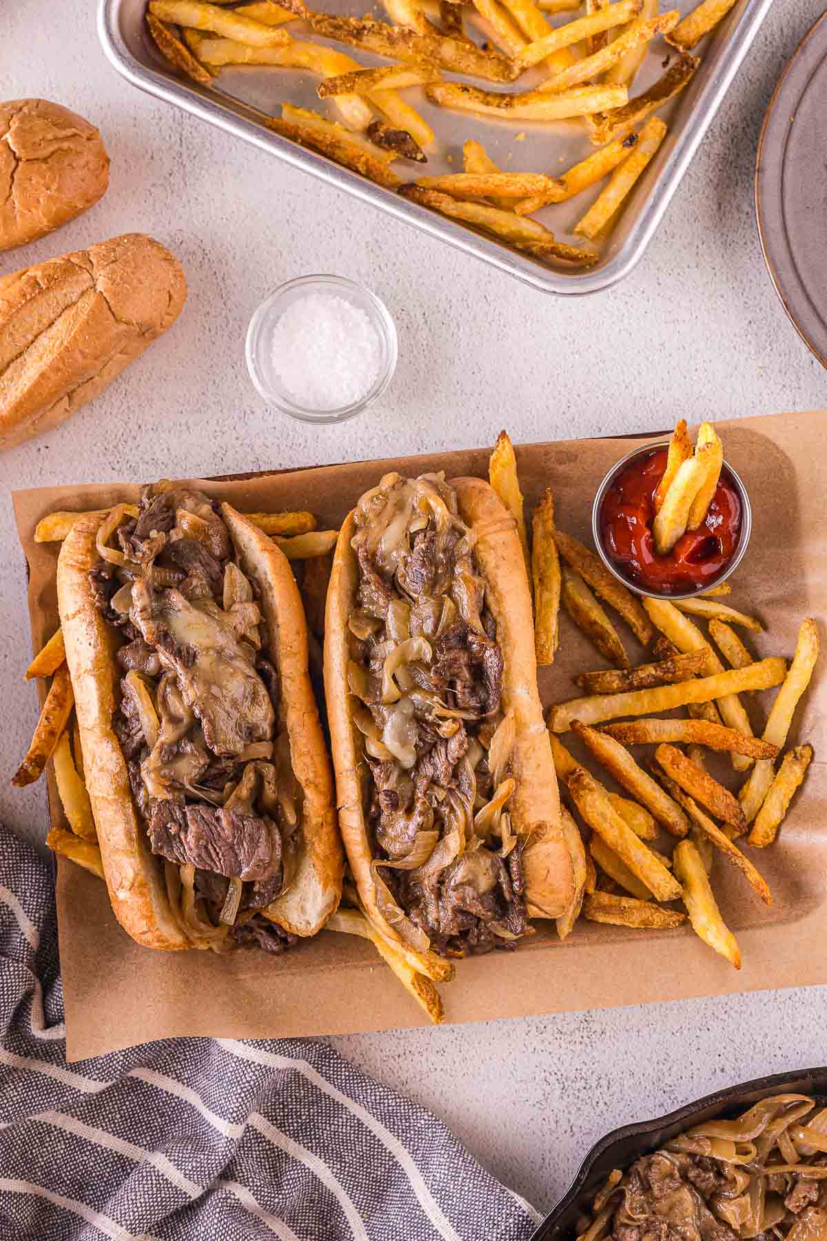 Dois sanduíches de cheesesteak com cebola salteada em uma bandeja, servidos com batatas fritas, uma xícara pequena de ketchup e um prato de sal.