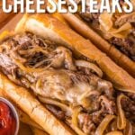 Dois sanduíches de cheesesteak Philly com queijo derretido, cebola salteada e carne fatiada em rolinhos hoagie, servidos em travessa de madeira com acompanhamento de ketchup.