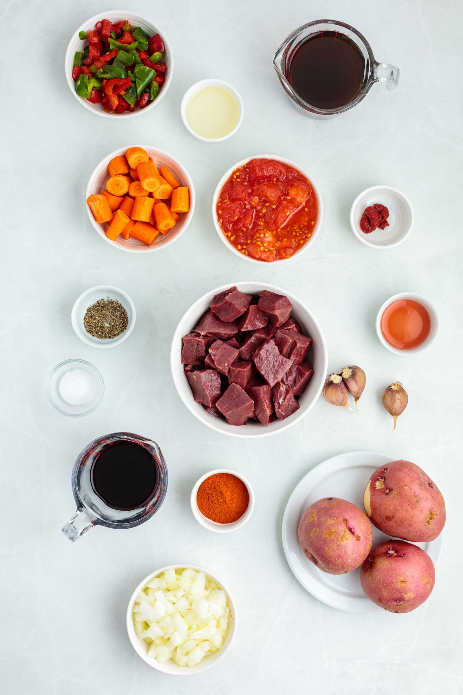 Vários ingredientes para um ensopado estão dispostos em uma superfície branca, incluindo coração de boi em cubos, batata, cebola, cenoura, alho, temperos, tomate, pimentão picado e líquidos em tigelas pequenas.