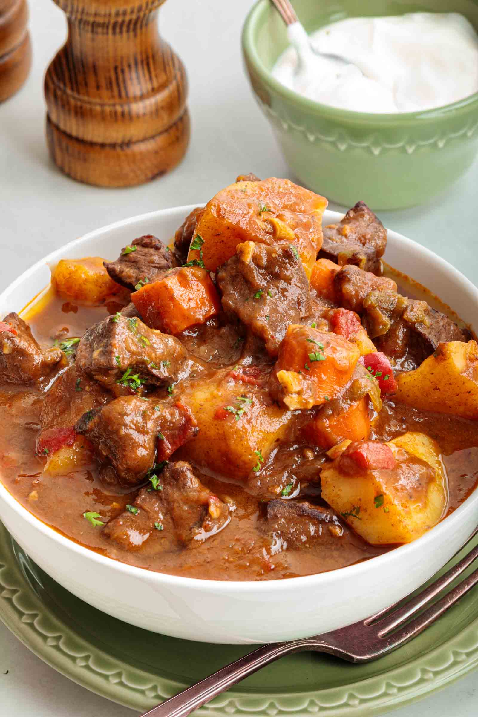 Tigela de ensopado de coração de boi com pedaços de carne, batatas e cenouras em molho marrom espesso, colocado em um prato com uma colher. Iogurte ou creme de leite e moinhos de pimenta ao fundo.