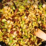Uma frigideira recheada com repolho frito e pedaços de bacon mexidos com uma espátula de madeira. Leituras de sobreposição de texto, "Como fazer repolho frito com bacon.