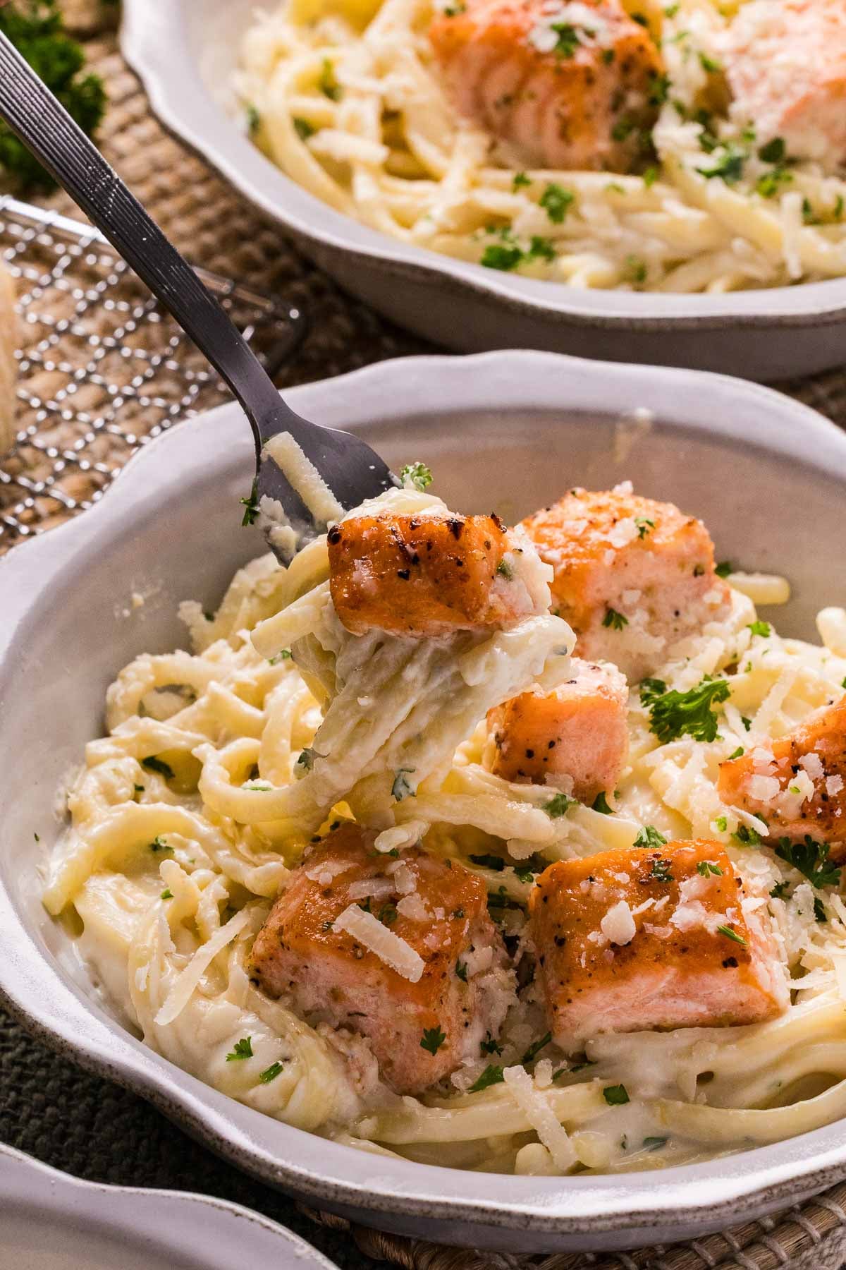 Uma tigela de salmão Alfredo, guarnecida com queijo ralado e salsa; um garfo contém um pedaço de macarrão e salmão.