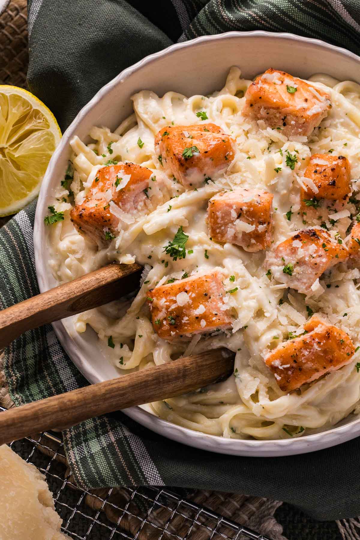 Uma tigela de macarrão linguini coberta com pedaços de salmão ao molho Alfredo, guarnecida com salsa e queijo ralado, com pinça de madeira e meio limão ao lado do prato.