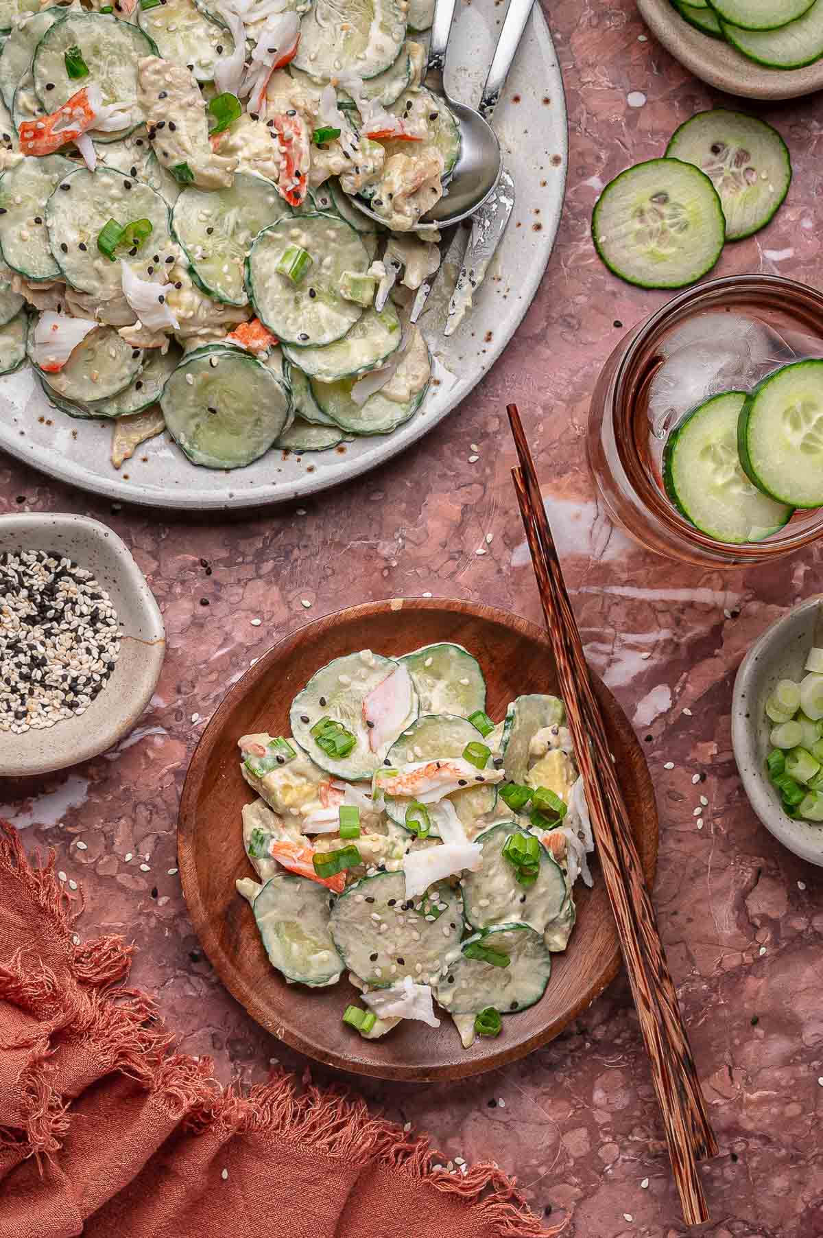 Uma pequena tigela de madeira com salada cremosa de pepino cali roll com palitos de caranguejo, guarnecida com cebolinha e sementes de gergelim, fica ao lado de um prato com a mesma salada e tigelas com rodelas de pepino e temperos.