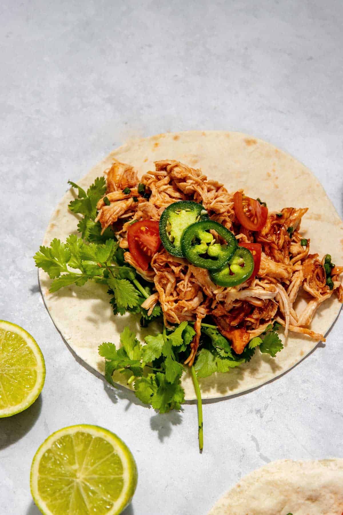 Um taco aberto recheado com frango desfiado, coentro, tomate cereja fatiado e jalapeños, apoiado sobre uma superfície cinza claro. Fatias de meio limão estão por perto.