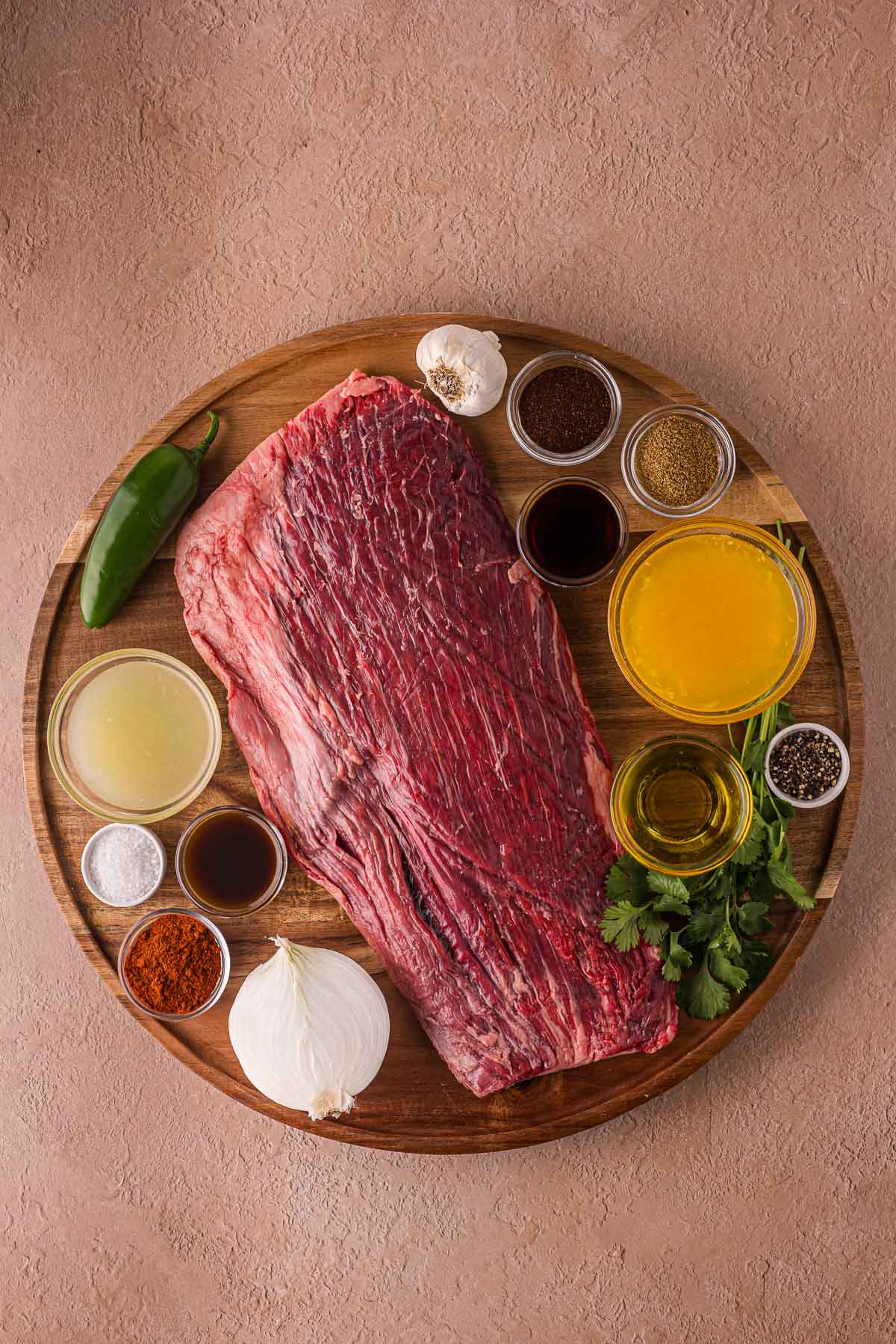 Bife de flanco cru em uma tábua de madeira, cercado por pequenas tigelas de temperos, molhos, caldo, ervas picadas, pimenta, bulbo de alho e cebola branca.