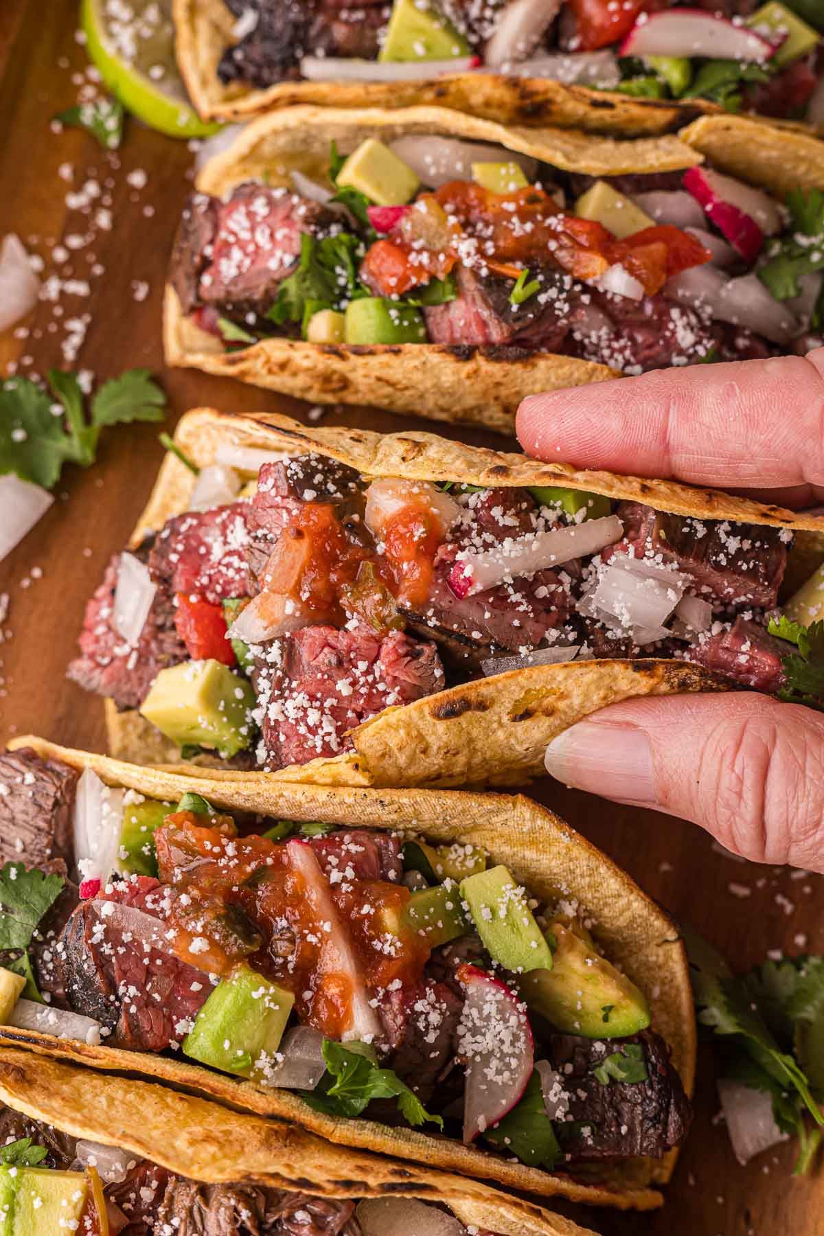 Uma mão segurando um taco de carne assada coberto com cebola em cubos, abacate, molho de tomate, rabanete e queijo esfarelado, ao lado de tacos semelhantes em uma superfície de madeira.