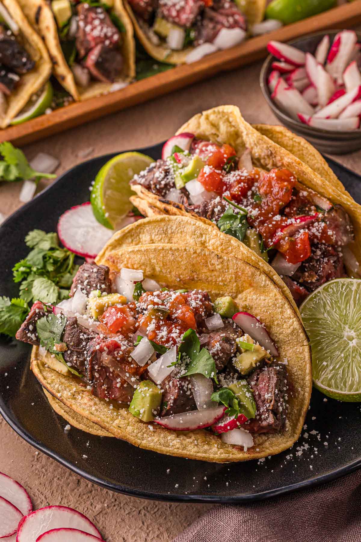 Dois tacos de carne assada em tortilhas de milho cobertas com cebola picada, coentro, molho, abacate, fatias de rabanete e queijo ralado, servidos com rodelas de limão em um prato preto.