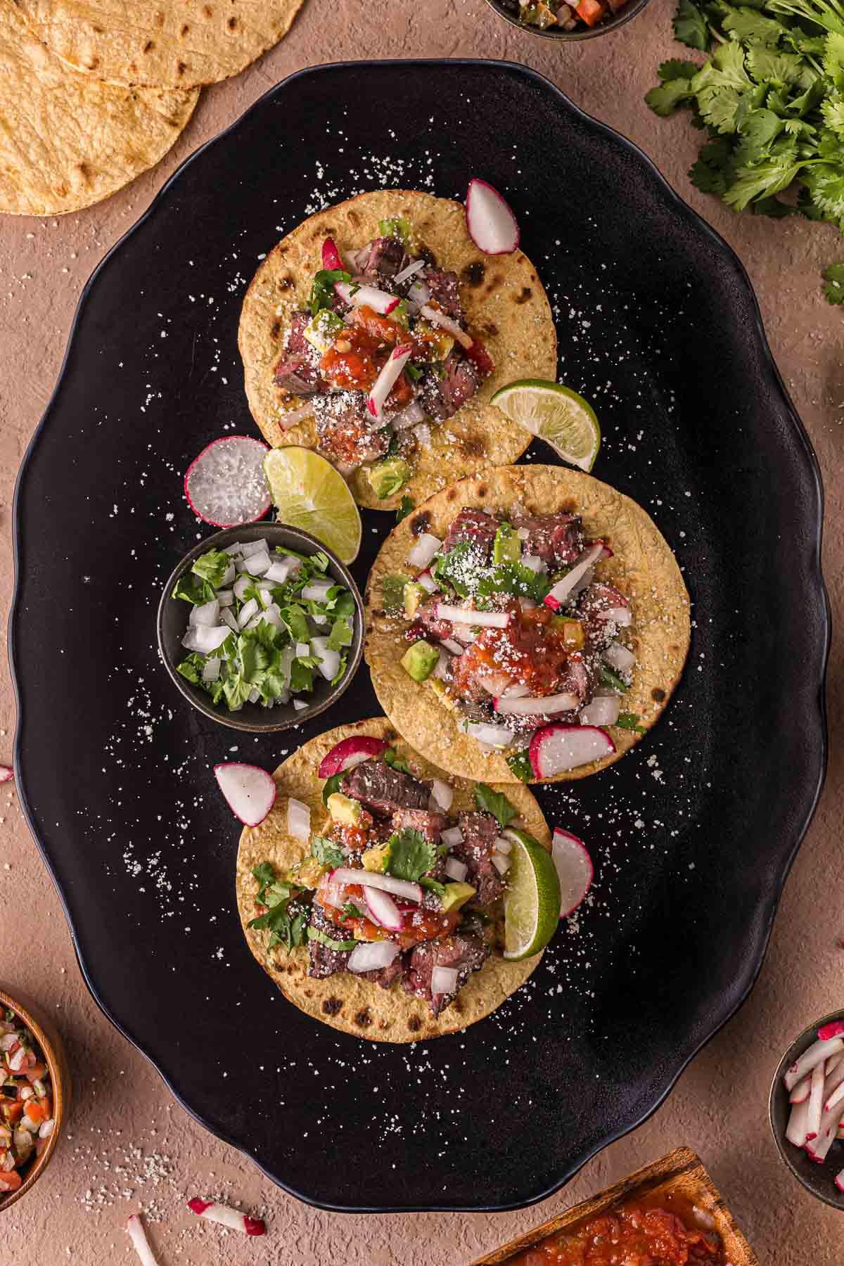 Três tacos de carne assada cobertos com cebola, coentro, rodelas de rabanete, molho e queijo, servidos com rodelas de limão e acompanhamento de cebola picada e coentro em uma travessa preta.
