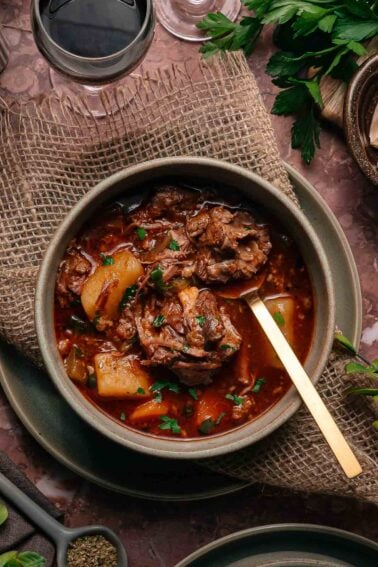 Uma tigela de guisado de rabada com batatas em caldo rico e escuro, guarnecido com ervas, colocado sobre uma esteira rústica com uma colher de ouro. Um copo de vinho tinto e ervas frescas estão por perto.