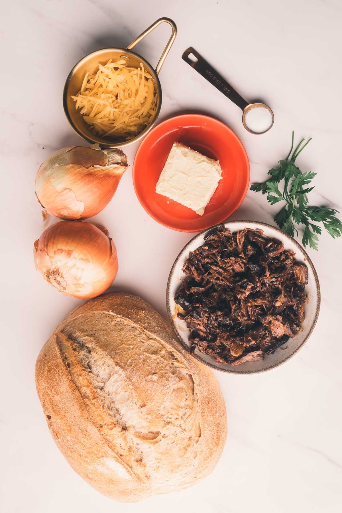 Um pão, duas cebolas, queijo ralado em um copo medidor, um quadrado de manteiga em um prato pequeno, salsa e uma tigela de carne desfiada sobre uma superfície branca.