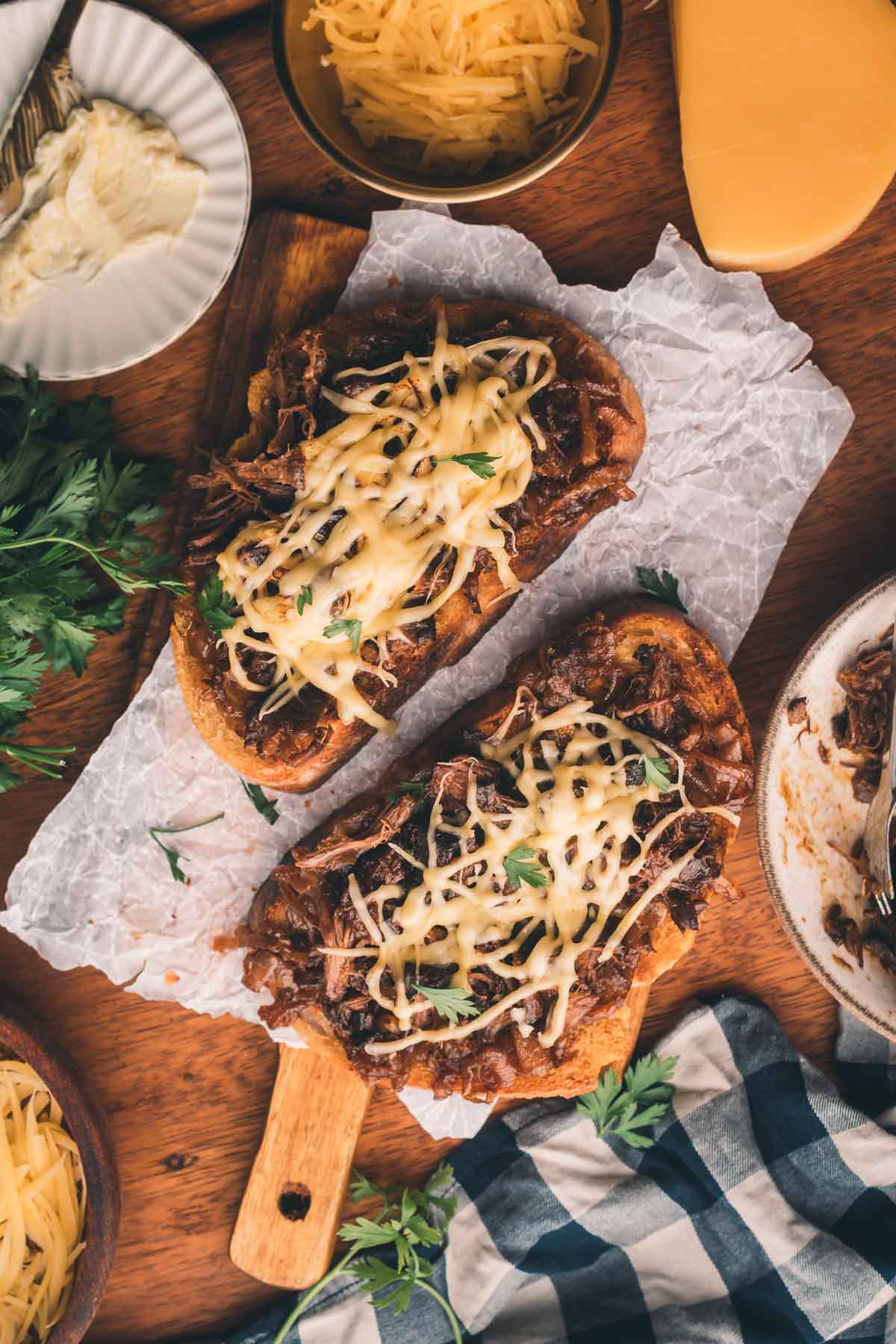 Dois sanduíches abertos cobertos com carne desfiada, queijo derretido e ervas frescas são colocados em papel manteiga, rodeados por tigelas de queijo ralado e salsa fresca.