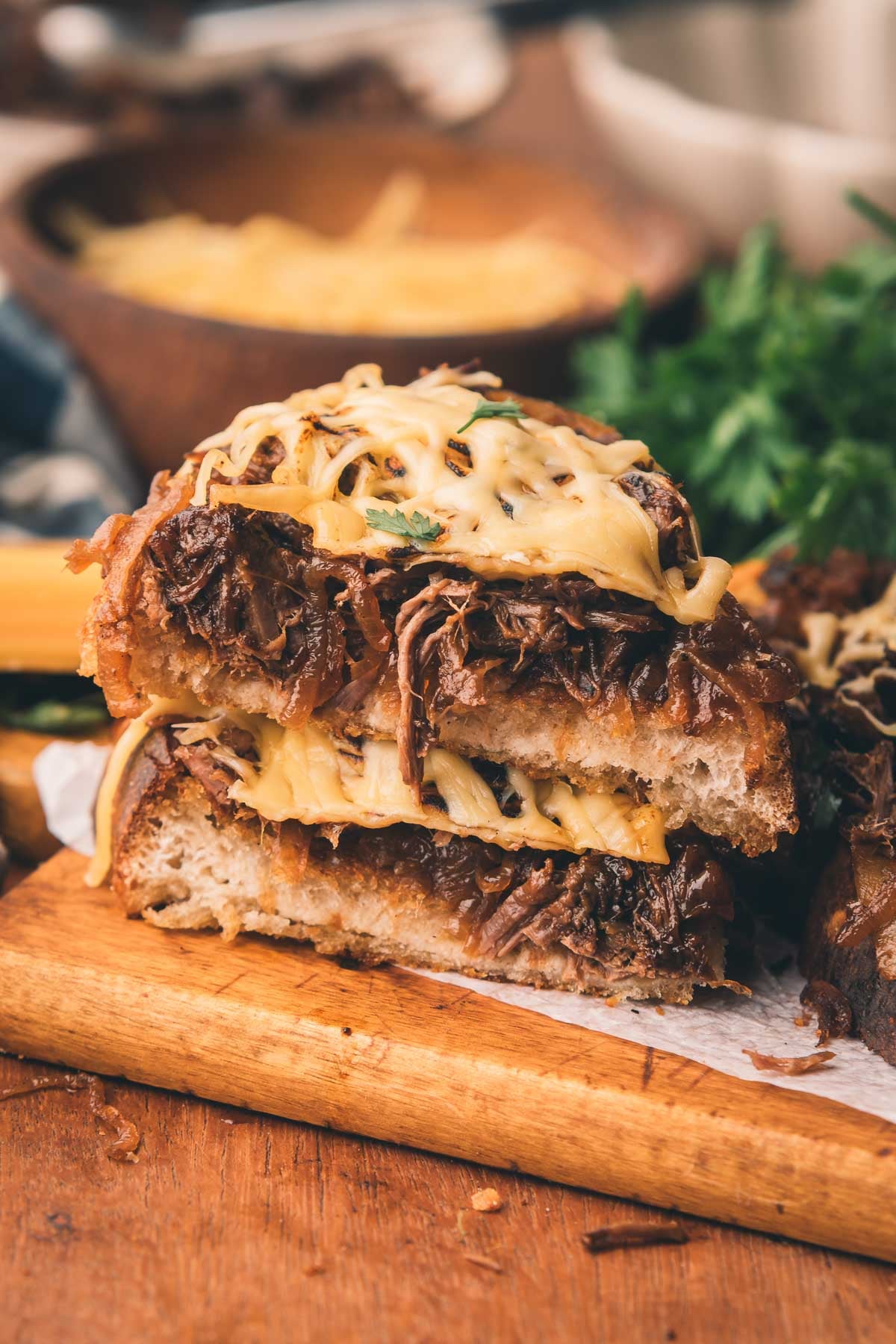 Close de um sanduíche de cara aberta empilhado com carne desfiada, queijo derretido e cebola caramelizada em pão torrado, servido em uma tábua de madeira.