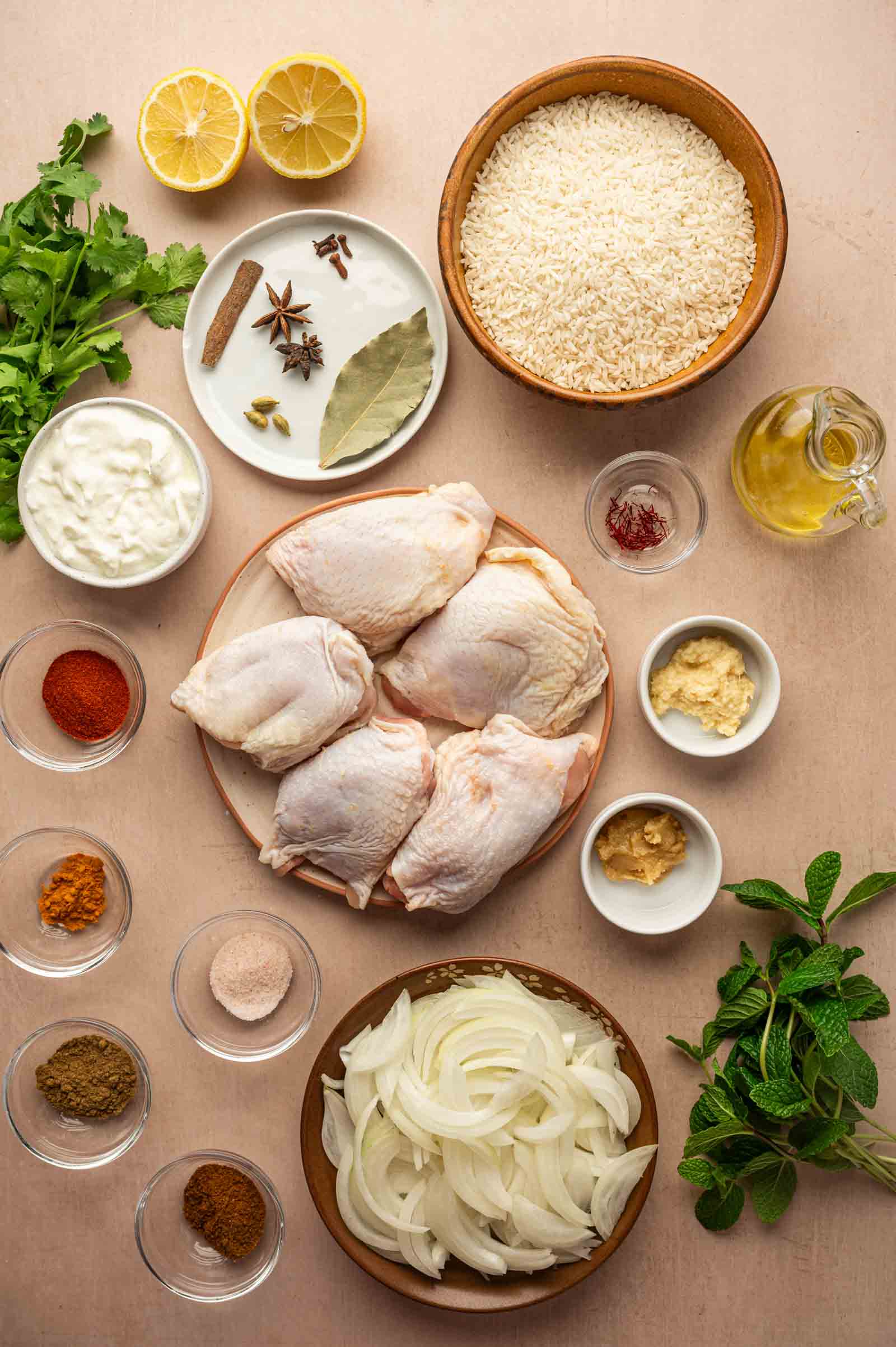Vista aérea de coxas de frango cru, tigelas de arroz, cebola fatiada, iogurte, especiarias, óleo, alho, gengibre, coentro fresco, hortelã e metades de limão dispostas sobre uma superfície leve.