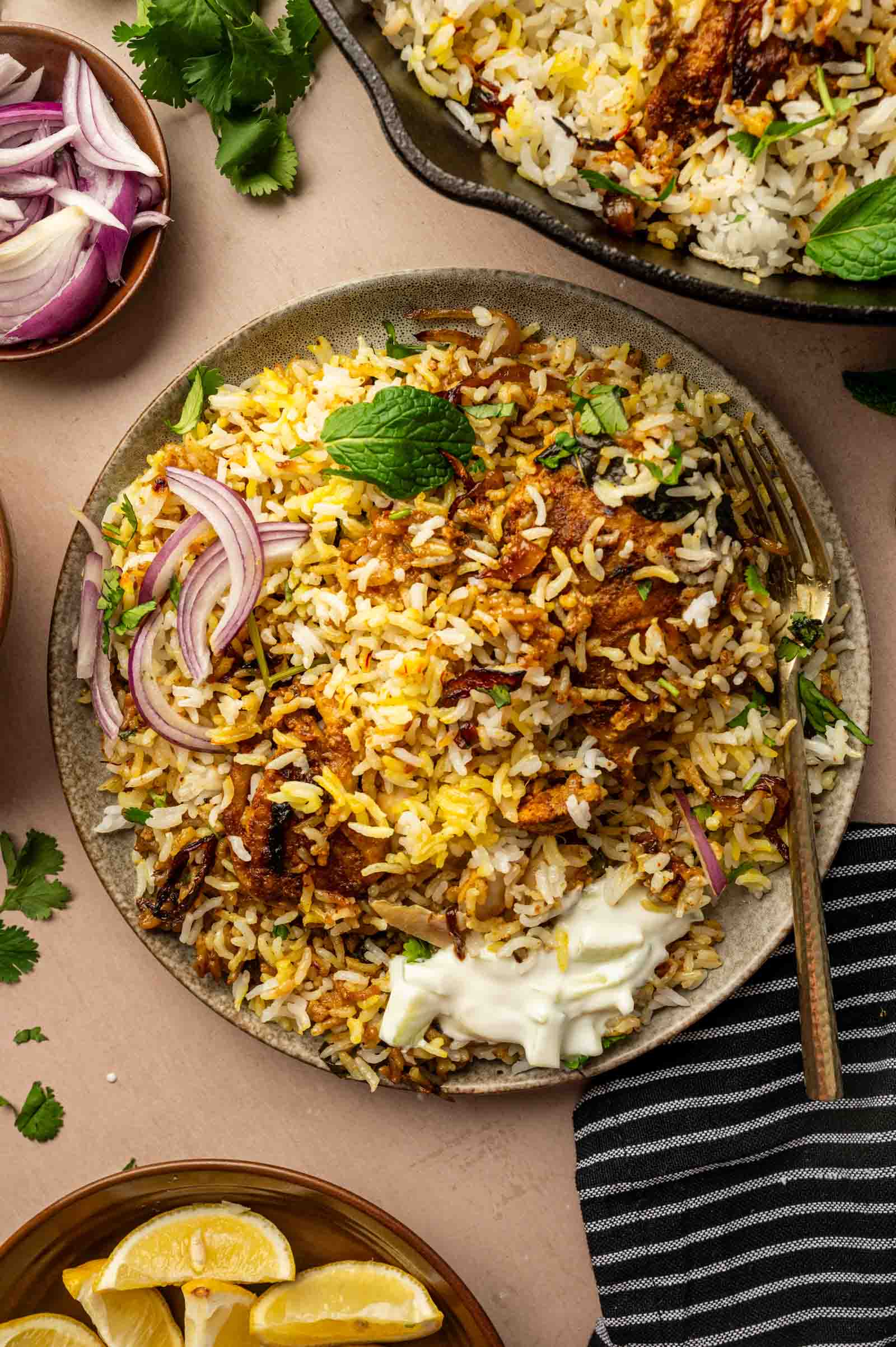 Um prato de arroz biryani de frango com pedaços de carne, guarnecido com rodelas de cebola roxa, ervas frescas, um bocado de iogurte e servido com rodelas de limão e cebola extra como acompanhamento.