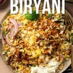 Um prato de frango biryani coberto com cebola roxa fatiada e ervas, com guardanapo listrado e garfo ao lado. Leituras de sobreposição de texto "Como fazer frango Biryani.