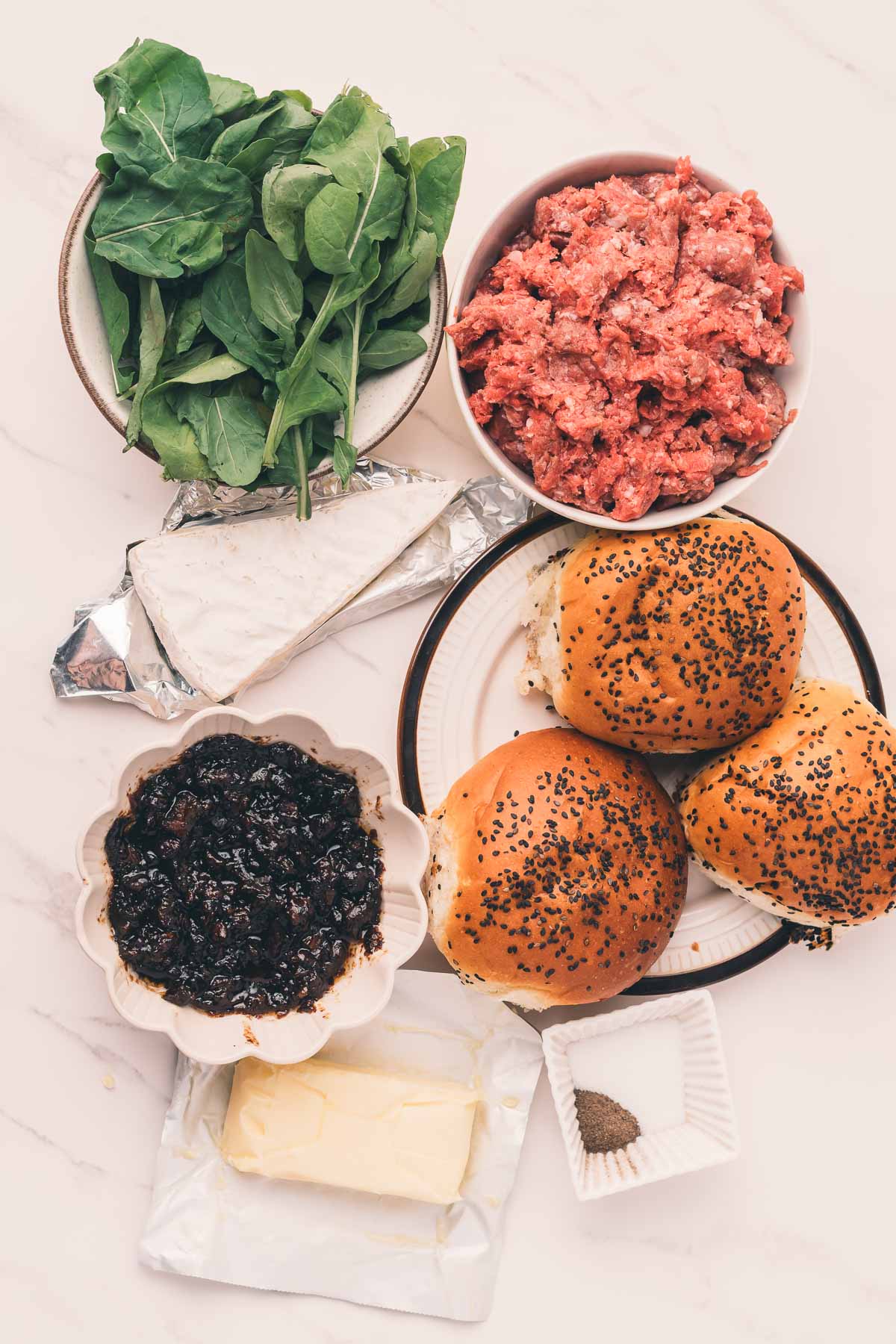 Vista de cima para baixo dos ingredientes do hambúrguer: carne moída, folhas verdes, queijo brie, manteiga, pães com sementes, geléia de bacon e um pequeno prato de pimenta preta.