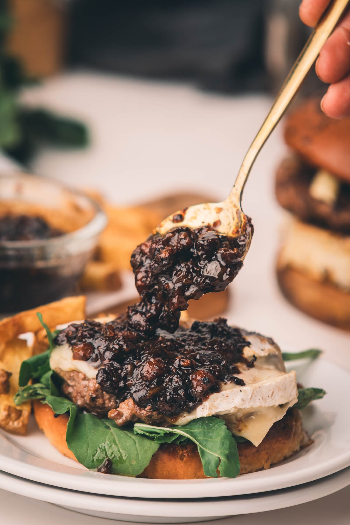 Uma mão coloca geléia de bacon escuro em um cheeseburger com verduras em um prato, com batatas fritas e outro hambúrguer ao fundo.